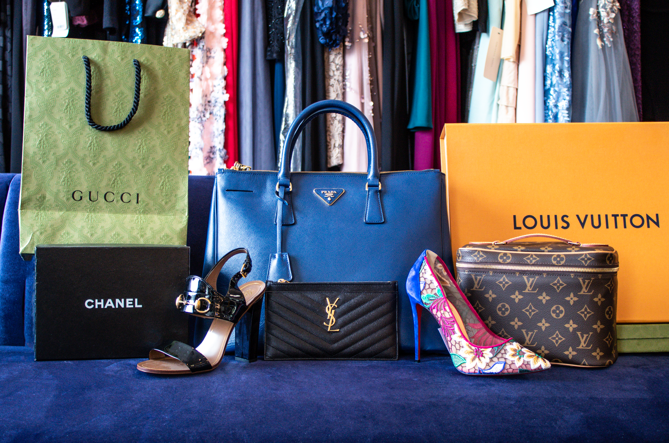 A photograph of a variety of handbags, shoes, and boxes/bags on a navy couch. The items on the couch, from left to right, are: Gucci Paper Bag, Chanel Box, Louboutin black heeled sandal, Navy Prada handle bag, Saint Laurent Clutch, Another Louboutin heel with pointed toe and decor, Louis Vuitton Makeup Pouch, and Louis Vuitton Box. Behind the couch is a rack with a variety of formal dresses as the background.