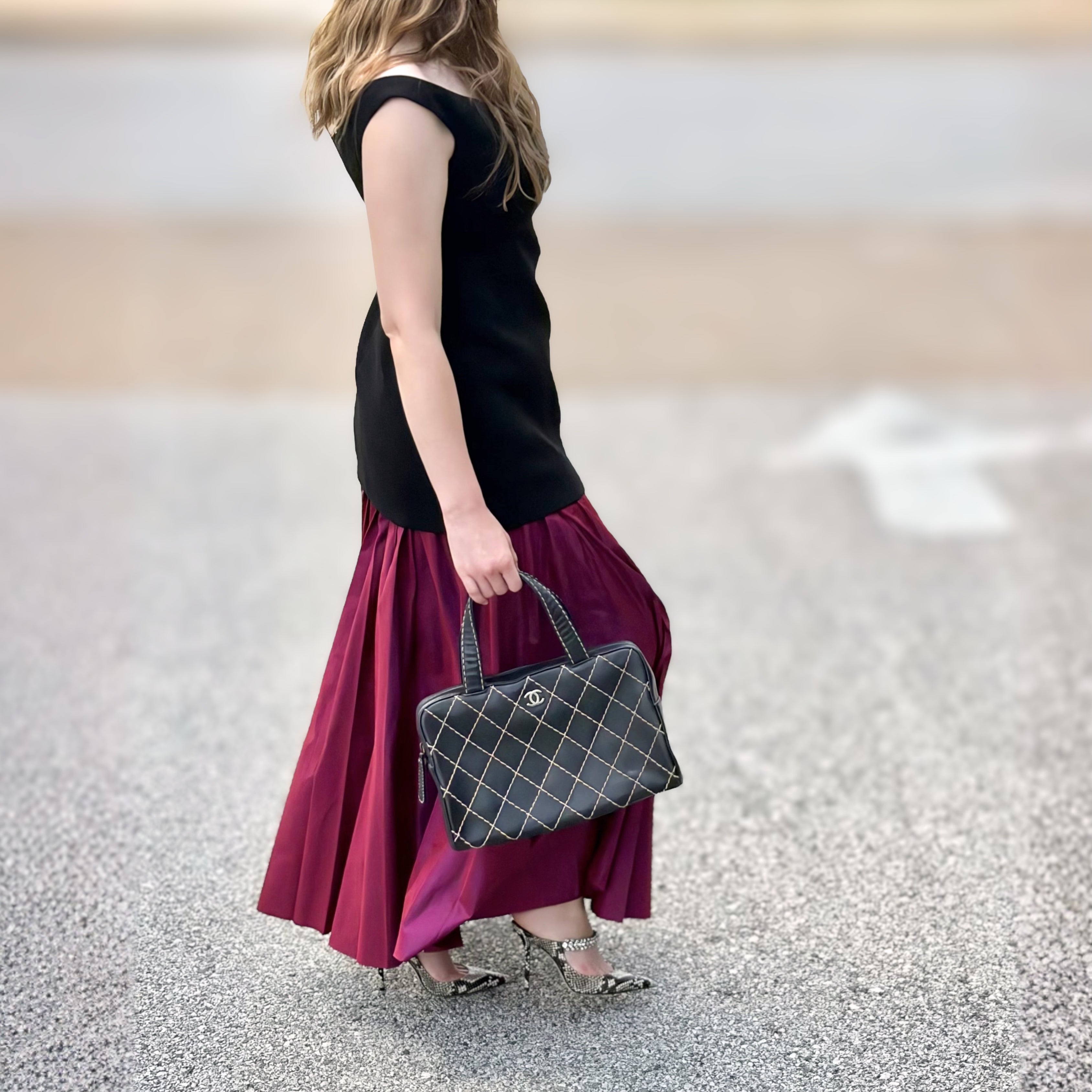 A woman from behind wearing a black sleeveless top and a pleated magenta skirt, carrying a black quilted handbag with a silver grid pattern and interlocking CC logo, walking on a paved surface.