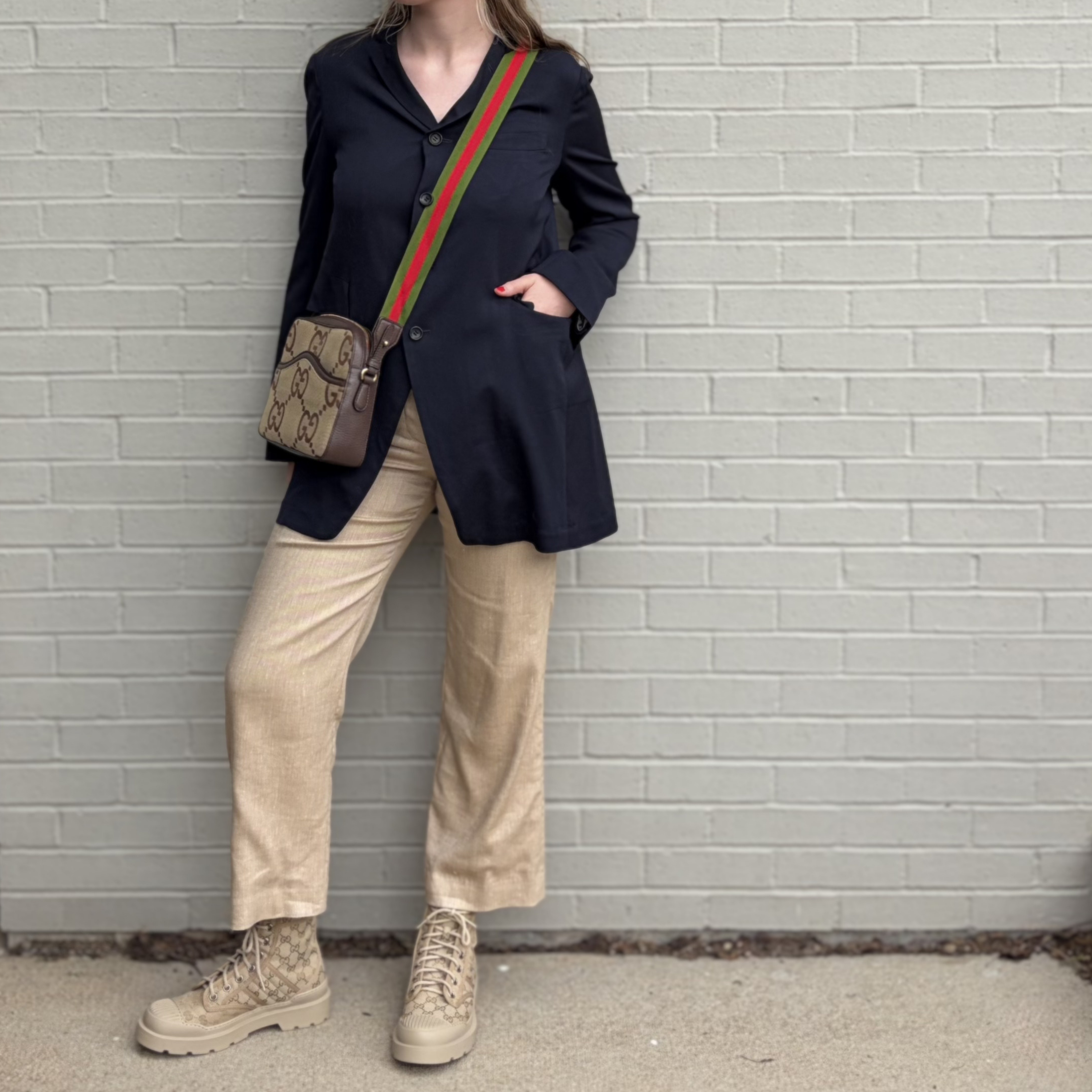 Person wearing a navy blazer, beige pants, and tan boots against a light gray brick wall.