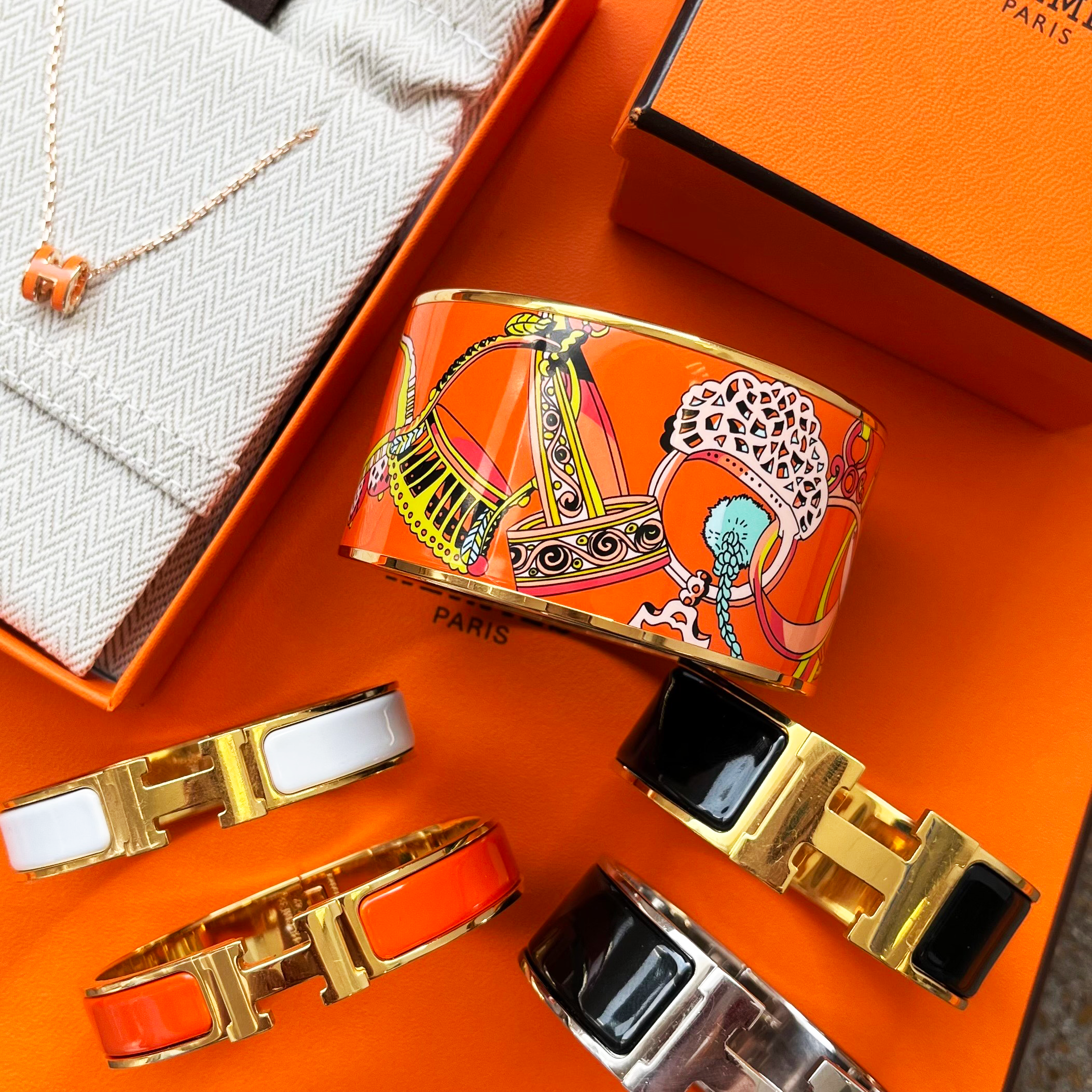 A set of Hermès H-curve cuff bracelets displayed on an orange Hermès box, alongside an orange and white branded pouch and a necklace. The bracelets feature orange, white, black, and silver enamel inlays with gold-tone metal.