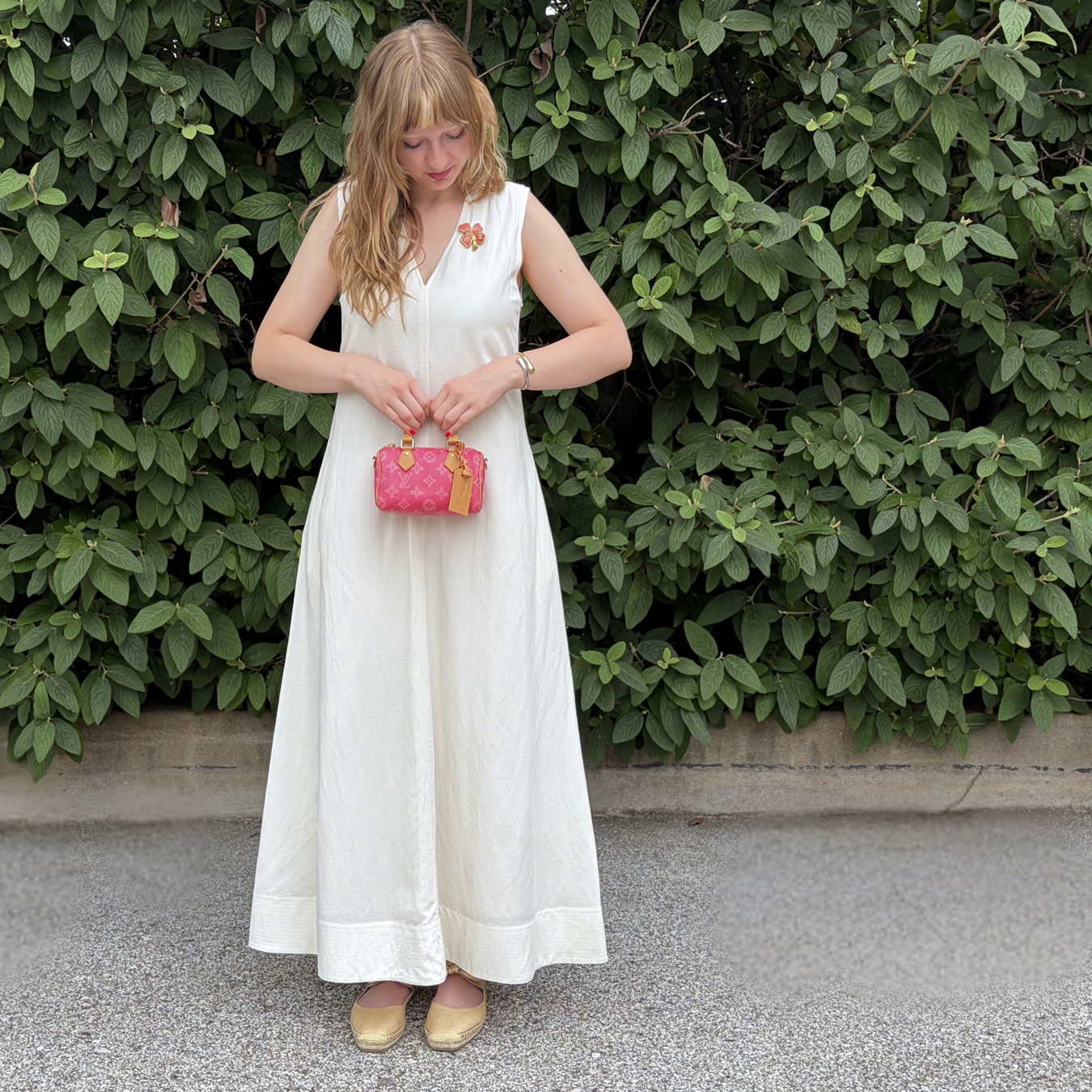 A woman in a white dress holding a pink Louis bag in front of foliage.