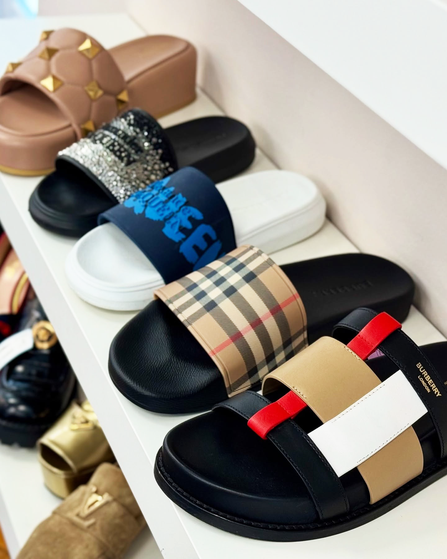 Collection of designer sandals in a variety of brands and colors on a white shelf.