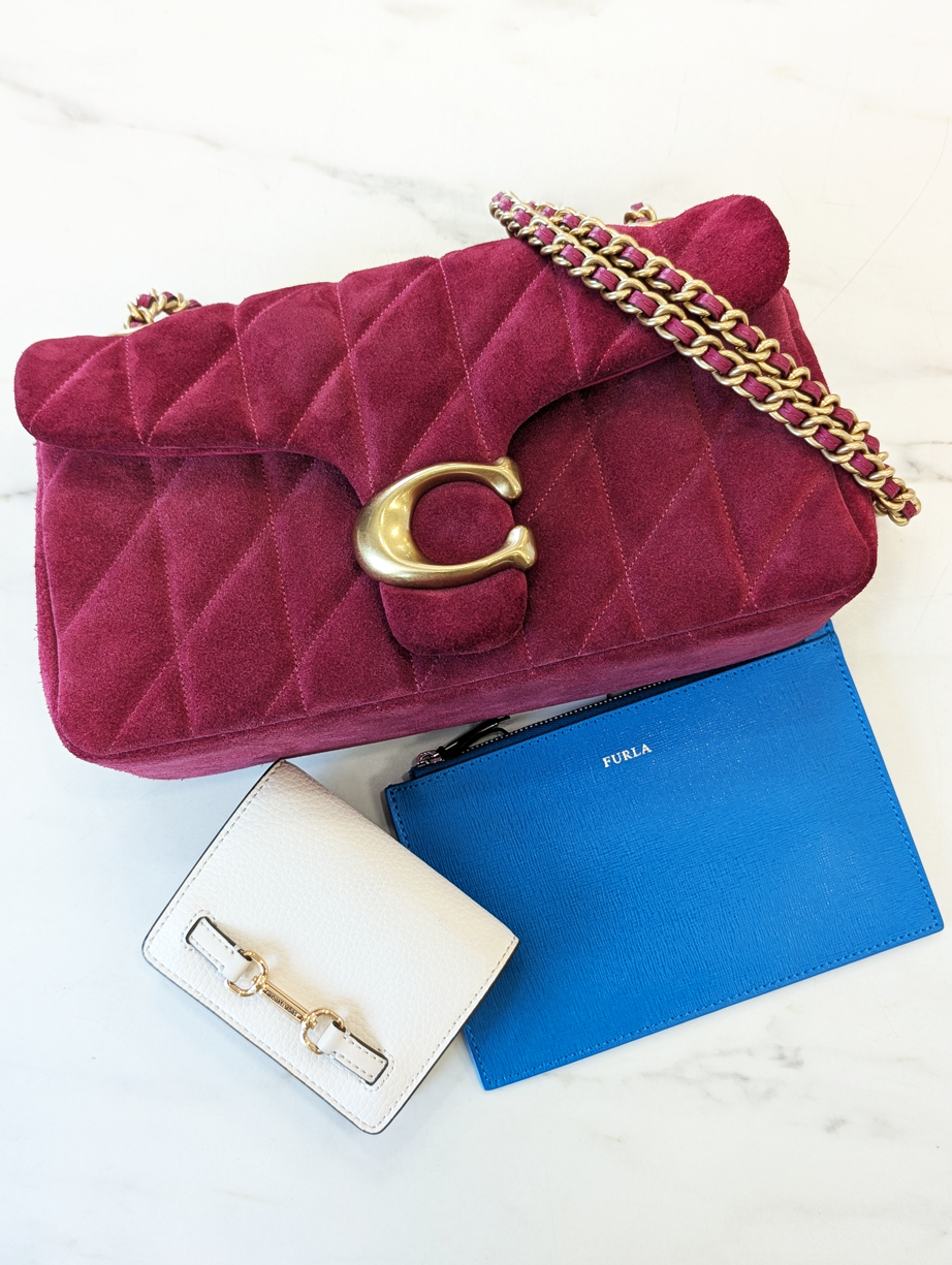 A burgundy Coach bag, a white Michael Kors wallet, and a blue Furla pouch on a white marbled background.