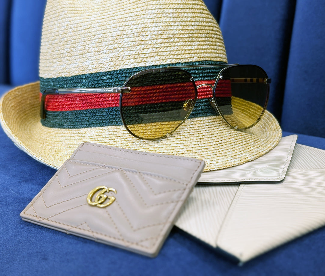A straw Gucci hat, Burberry sunglasses, and a Gucci and Louis Vuitton wallet on a navy background.