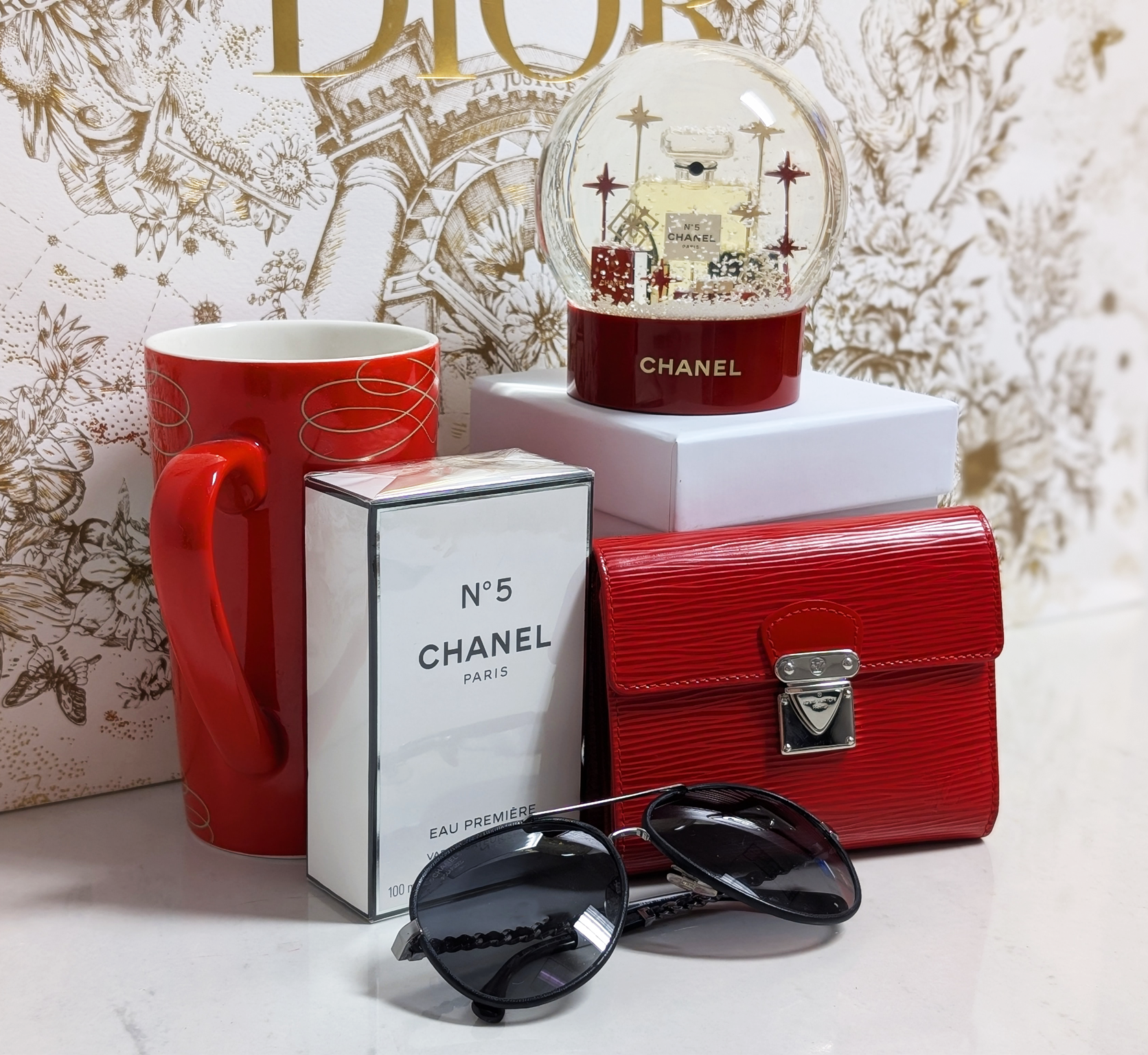 A Chanel Snow Globe, Perfume, and sunglasses, as well as a red epi leather Louis Vuitton wallet and a holiday-themed mug against a gold and white background.