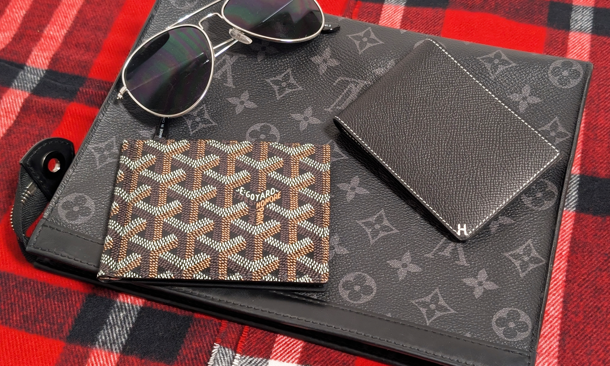 A photo of a Louis Vuitton pouch, an Hermès men's bifold wallet, a Goyard men's bifold wallet, and a pair of aviator sunglasses on top of two red, black and white plaid scarves.