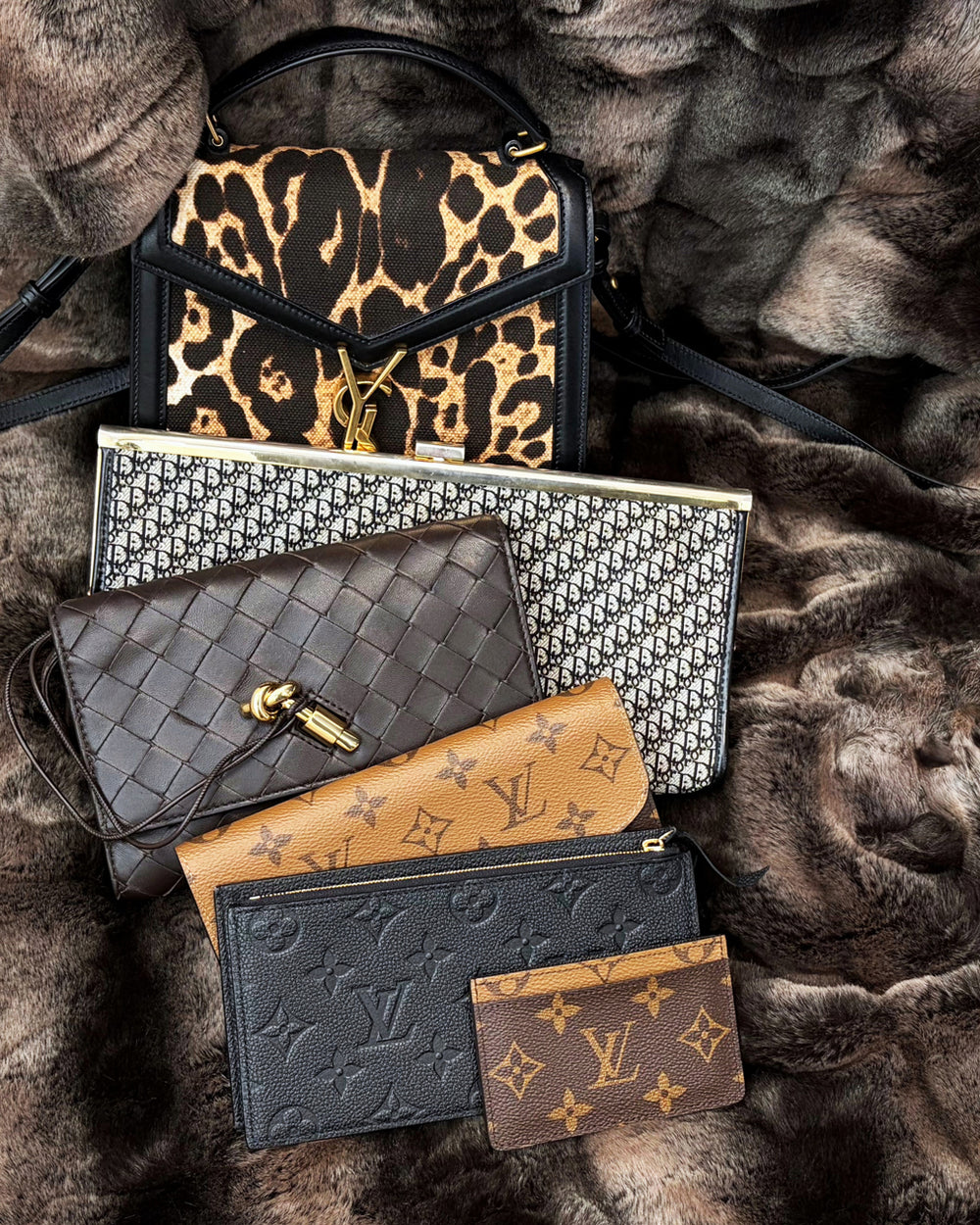 A curated flat-lay of luxury designer accessories arranged on a brown fur background, including a leopard-print shoulder bag, a patterned clutch, a woven black pouch, a tan monogram wallet, a dark monogram card holder, and a small brown monogram card.