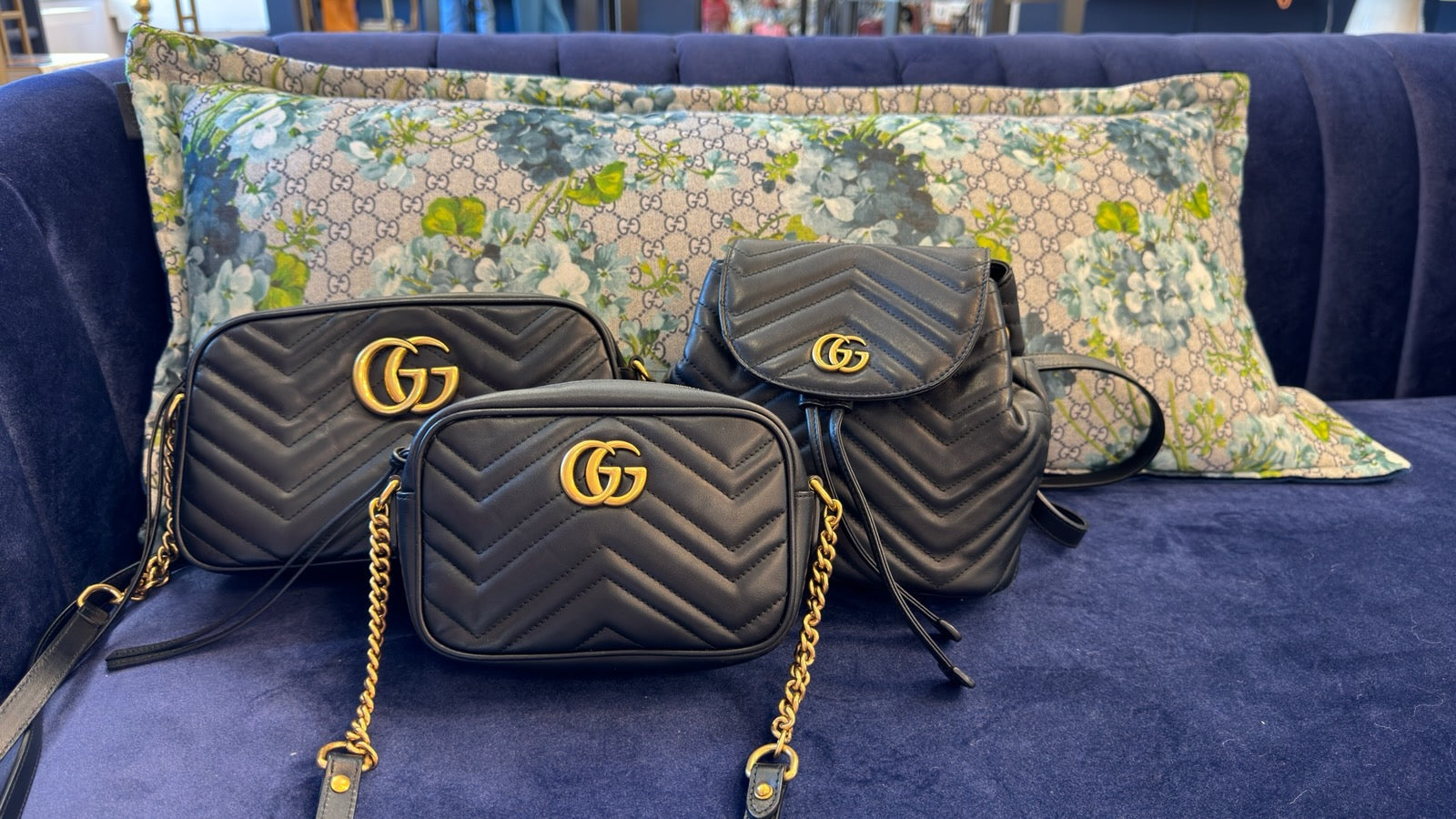 An image of 3 black Gucci marmont bags (two crossbodies and a backpack) on a navy couch. There is a Gucci pillow behind them with the signature Gucci GG monogram, as well as a blue floral painted pattern.