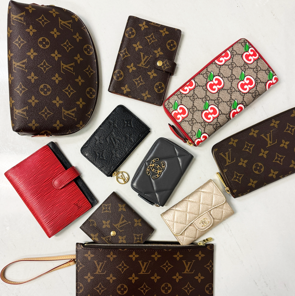 A curated flat-lay collection of luxury handbags and wallets, featuring prominent Louis Vuitton monogram canvas, a red textured leather wallet, a black embossed leather wallet, a black quilted wallet with a gold-tone logo, a beige quilted wallet, and a Gucci monogram wallet with red and green cherry pattern.