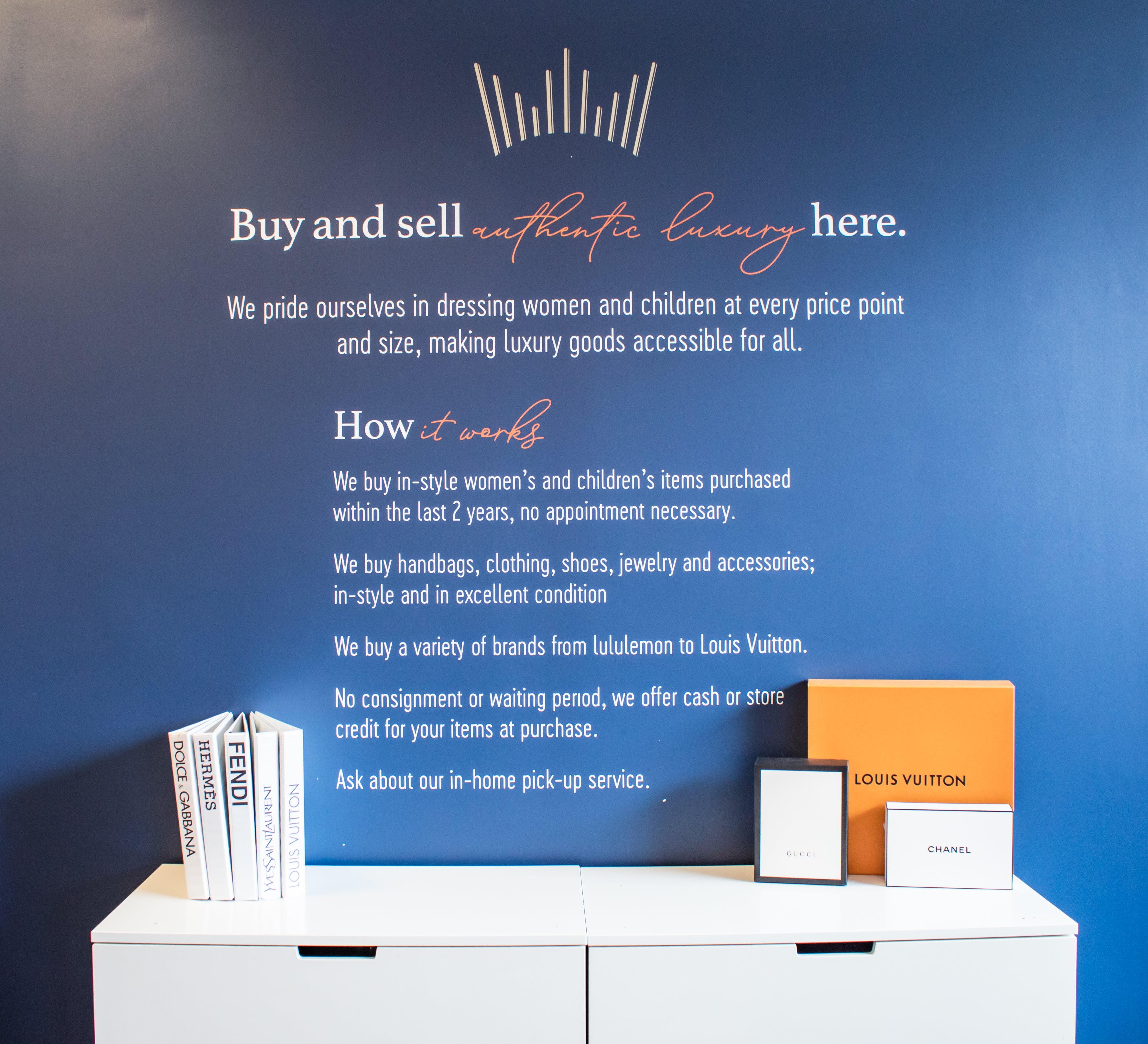 Blue wall with text about buying and selling authentic luxury items, featuring brand boxes on a white surface.